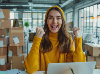 Junge Frau in gelbem Pullover jubelt vor Laptop inmitten eines Logistikbüros voller Pakete – Ausdruck von Erfolg und Begeisterung